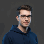 Porträt von Stephan Depta, jungem Mann mit kurzem, dunklem Haar und Brille. Er trägt einen dunkelblauen Hoodie und blickt freundlich in die Kamera. Der Hintergrund ist grau und weich ausgeleuchtet, wodurch das Gesicht klar betont wird.