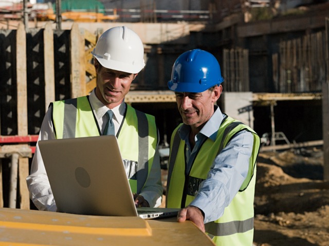 Zwei Bauingenieure oder Architekten in Warnwesten und Schutzhelmen (einer weiß, einer blau) stehen auf einer sonnigen Baustelle und betrachten gemeinsam konzentriert einen Laptop. Sie stehen vor einem Rohbau mit Holzverschalungen und Betonstrukturen.